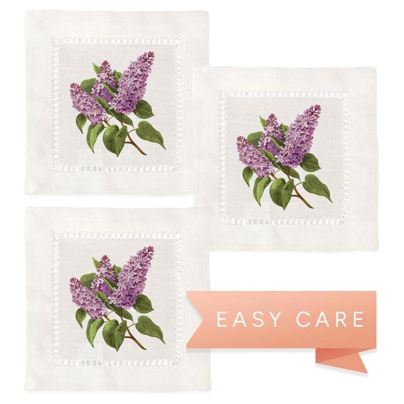 May include: Set of three white linen cocktail napkins with a lilac floral design. Each napkin features a cluster of purple lilacs with green leaves. The napkins have a scalloped edge and are machine washable.