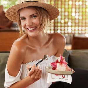 May include: A woman wearing a straw hat and a white off-the-shoulder top is holding a spoon and a small plate with a pink and white dessert. The dessert is topped with pink flowers and a red sauce.