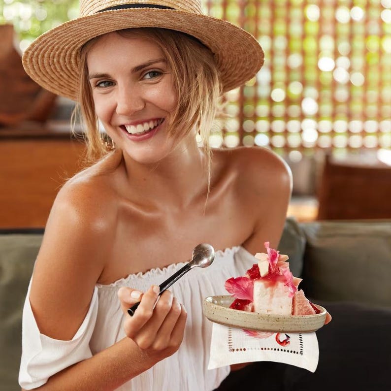May include: A woman wearing a straw hat and a white off-the-shoulder top is holding a spoon and a small plate with a pink and white dessert. The dessert is topped with pink flowers and a red sauce.