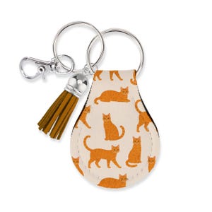 May include: A teardrop-shaped keychain featuring a repeating pattern of orange tabby cats on a cream background. The keychain includes a silver-toned metal split ring, a lobster clasp, and a short, brown faux leather tassel attached to the top.
