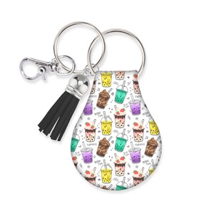 May include: A white keychain with a pattern of colorful boba tea drinks. The keychain has a silver ring, a black tassel, and a lobster clasp. The boba tea cups are in various colors, with the words "BOBA TEA" and "YUMMY!" printed on them.