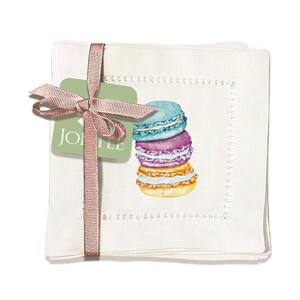 Puede incluir: Juego de cuatro servilletas de lino blanco con una ilustración de acuarela de macarons coloridos. Las servilletas tienen un delicado borde bordado y están empaquetadas con una etiqueta verde que dice "Jolie".