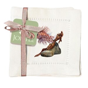 May include: Set of four white linen napkins with a red and white lionfish illustration on a brown driftwood and gray rock background. The napkins are tied with a pink ribbon and a green tag that says "JORTIE".