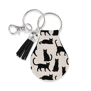May include: A teardrop-shaped keychain featuring a repeating pattern of black cats on a light beige background. The keychain includes a silver-toned metal split ring, a lobster clasp, and a black faux leather tassel attached to the top hardware.
