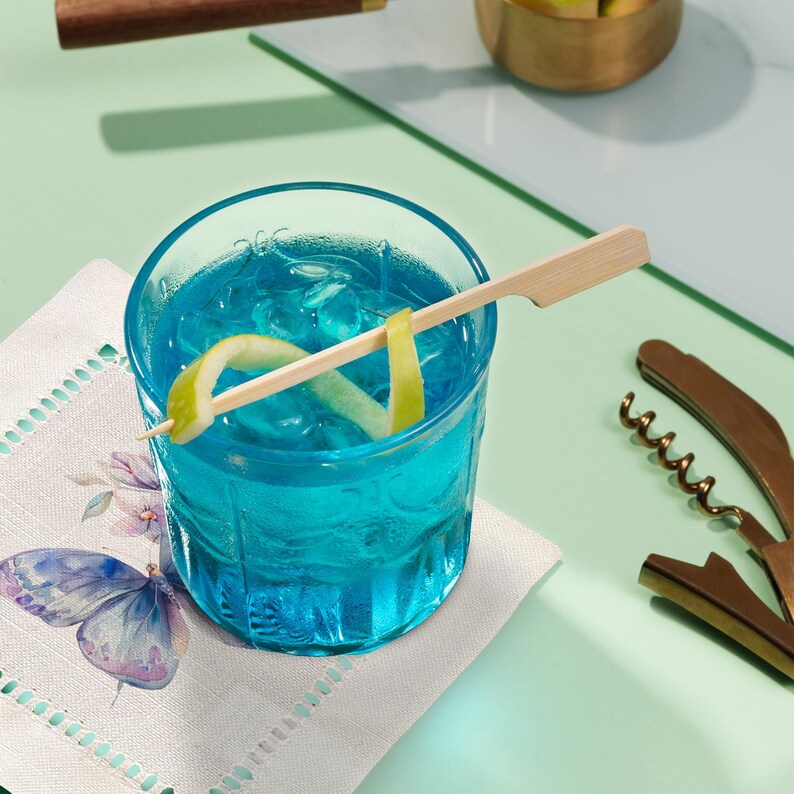 K&ouml;nnte beinhalten: Ein blauer Cocktail mit Eis und einer Zitronenzeste, garniert mit einem Holzspie&szlig; in einem klaren Glas auf einer wei&szlig;en Serviette mit einem Schmetterlingsmuster.