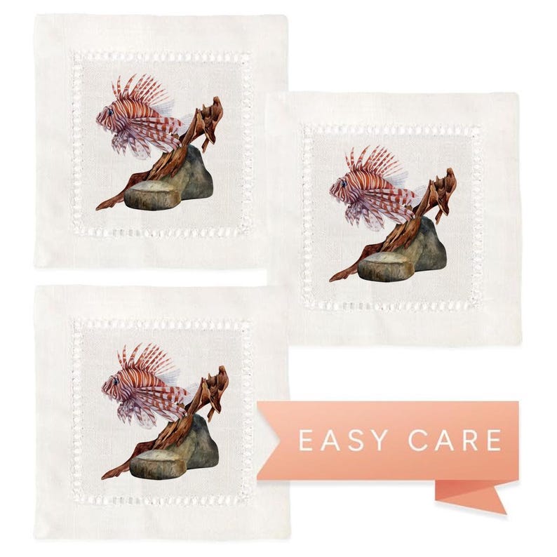 May include: Set of three linen cocktail napkins featuring a watercolor illustration of a lionfish on a piece of driftwood. The napkins have a white background with a white scalloped border.