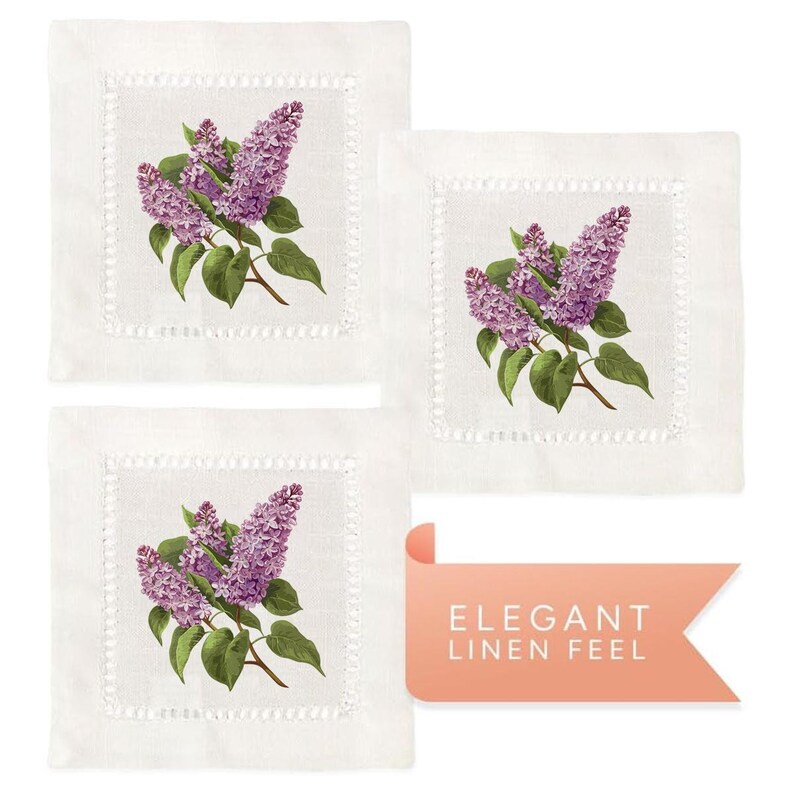 May include: Set of three white linen cocktail napkins with a delicate embroidered border. Each napkin features a vintage illustration of a sprig of purple lilac flowers.