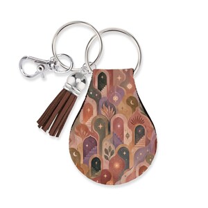 May include: A teardrop-shaped keychain featuring a repeating pattern of arched doorways, staircases, stars, and crescent moons in shades of terracotta, mauve, and sage green. It includes a silver metal split ring, a lobster clasp, and a brown tassel.
