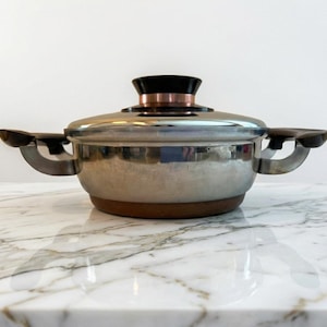 May include: A stainless steel cooking pot with a lid. The pot has a brown base and handles, with a black and copper-colored knob on the lid. The pot is sitting on a marble surface.