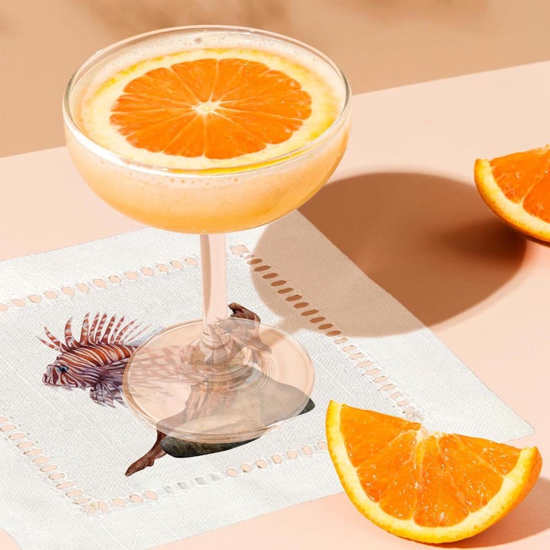 May include: A cocktail in a coupe glass with a slice of orange on top. The glass is sitting on a white napkin with a seahorse design. There are two orange slices on the table next to the glass.