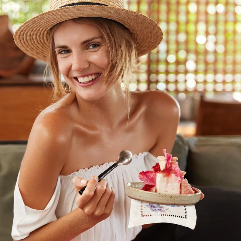 K&ouml;nnte beinhalten: Eine Frau mit einem Strohhut l&auml;chelt, w&auml;hrend sie einen L&ouml;ffel und einen kleinen Teller mit einem rosa und wei&szlig;en Dessert h&auml;lt, das mit Fr&uuml;chten garniert ist.