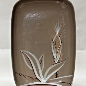 May include: A rectangular, brown glass dish with rounded corners. The dish features a white and brown floral design on the front. The design includes stylized leaves and a central flower, adding a touch of elegance.