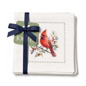 May include: A stack of square off-white fabric napkins featuring a printed illustration of a red cardinal bird perched on a branch with green leaves and red berries. The napkins are tied with a dark blue ribbon and a green tag labeled JOI TEE.