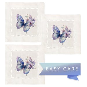 K&ouml;nnte beinhalten: Set aus drei wei&szlig;en Leinen-Servietten mit einer zarten, gestickten Bord&uuml;re. Jede Serviette zeigt ein Aquarell-Design eines blauen Schmetterlings mit lila Blumen.