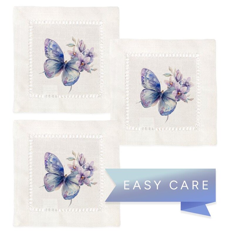 K&ouml;nnte beinhalten: Set aus drei wei&szlig;en Leinen-Servietten mit einer zarten, gestickten Bord&uuml;re. Jede Serviette zeigt ein Aquarell-Design eines blauen Schmetterlings mit lila Blumen.