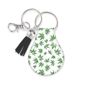 May include: A white leather keychain with a green marijuana leaf pattern. The keychain has a silver key ring and a black tassel.