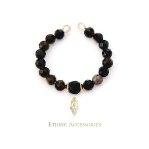 Può includere: Un anello di perline nere con dettagli dorati. L'anello è composto da perline nere sfaccettate e una perlina centrale dorata. Le parole "Ethnic Accessories" sono stampate sotto.