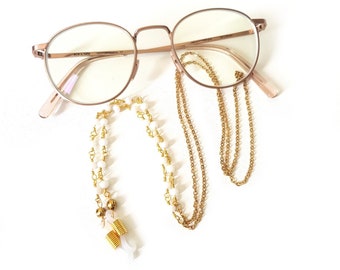 Könnte beinhalten: Eine runde Brille mit goldfarbenem Rahmen und klaren Gläsern sowie zwei Brillenketten. Eine Kette ist golden, die andere weiß mit goldenen Akzenten.