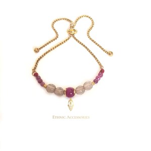 May include: A gold chain bracelet with pink and gray beads and a gold pendant with a face design. The bracelet has an adjustable sliding clasp. "ETHNIC ACCESSORIES" is printed below the bracelet.