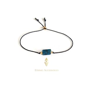 May include: A simple adjustable bracelet with a dark blue gemstone bead and gold accents. The bracelet is made with black cord and has a sliding knot closure.
