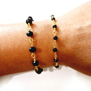 May include: A delicate bracelet featuring black faceted beads strung on gold-colored wire. The beads vary in size and are evenly spaced along the wire, creating a simple yet elegant design. The bracelet is shown on a wrist.