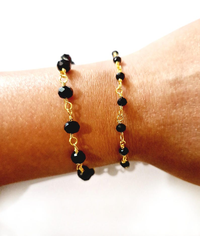 May include: A delicate bracelet featuring black faceted beads strung on gold-colored wire. The beads vary in size and are evenly spaced along the wire, creating a simple yet elegant design. The bracelet is shown on a wrist.
