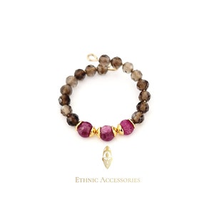 Op de afbeelding: Een kralenring met rookkwarts en robijn edelstenen. De ring heeft een goudkleurige draadband en is versierd met drie robijnkralen. De tekst "Ethnic Accessories" staat eronder.