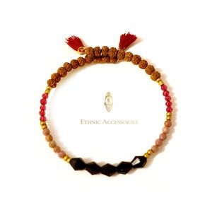 Puede incluir: Un brazalete de cuentas multicolores con borlas rojas en cada extremo. El brazalete presenta cuentas marrones, rosas y rojas, así como cuentas facetadas negras. El texto "ETHNIC ACCESSORIES" es visible en el fondo.