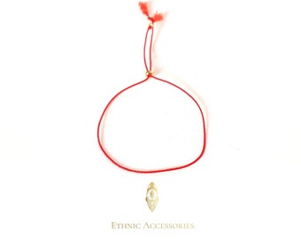 May include: Red string bracelet with a gold charm featuring an African mask design. The bracelet is tied with a knot at the end.