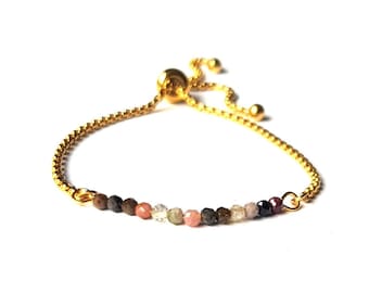 May include: A gold-toned adjustable bracelet with a delicate chain and a row of colorful, faceted beads. The beads include shades of brown, pink, and clear, creating a subtle, elegant design. The bracelet is set against a white background.
