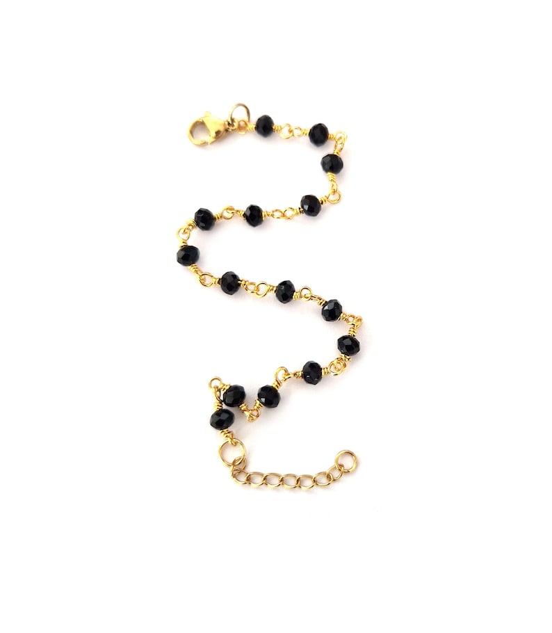 May include: A delicate bracelet featuring black faceted beads strung on a gold-colored chain. The bracelet has a lobster clasp closure and an adjustable chain for a customizable fit. The beads are evenly spaced along the chain.