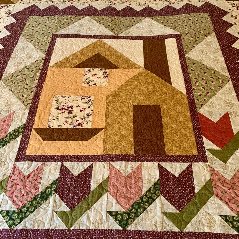 K&ouml;nnte beinhalten: Eine Quiltdecke mit einem braunen und braunen Hausdesign in der Mitte. Das Haus ist von einem Rand aus gr&uuml;nem, braunem und rosafarbenem Blumenstoff umgeben. Die Quiltdecke ist mit einem Wirbelmuster gesteppt.