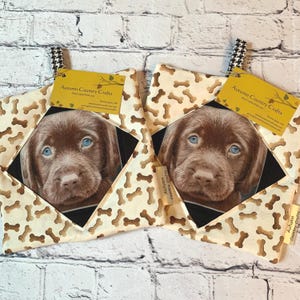 May include: Two square pot holders with a brown puppy with blue eyes. The pot holders have a cream background with brown bone shapes. Each pot holder has a black and white patterned hanging loop and a tag that reads "Autumn Country Crafts."