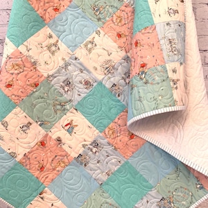 May include: A patchwork quilt featuring alternating squares of solid teal fabric and printed fabric with Peter Rabbit illustrations in shades of pink, blue, and cream. The quilt has a white backing with a swirling quilted pattern and striped binding.