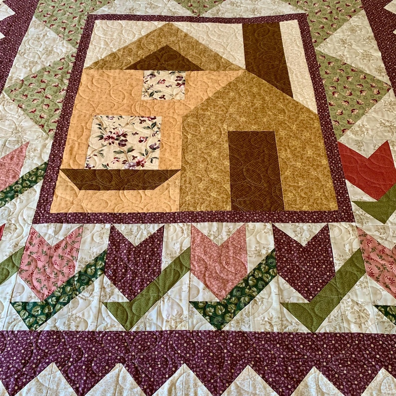 K&ouml;nnte beinhalten: Eine Steppdecke mit einem braunen und braunen Hausdesign in der Mitte. Die Steppdecke ist aus verschiedenen Stoffen gefertigt, darunter Blumenmuster und Uni-Farben. Die Steppdecke ist mit einem burgunderfarbenen und wei&szlig;en Rand eingefasst.