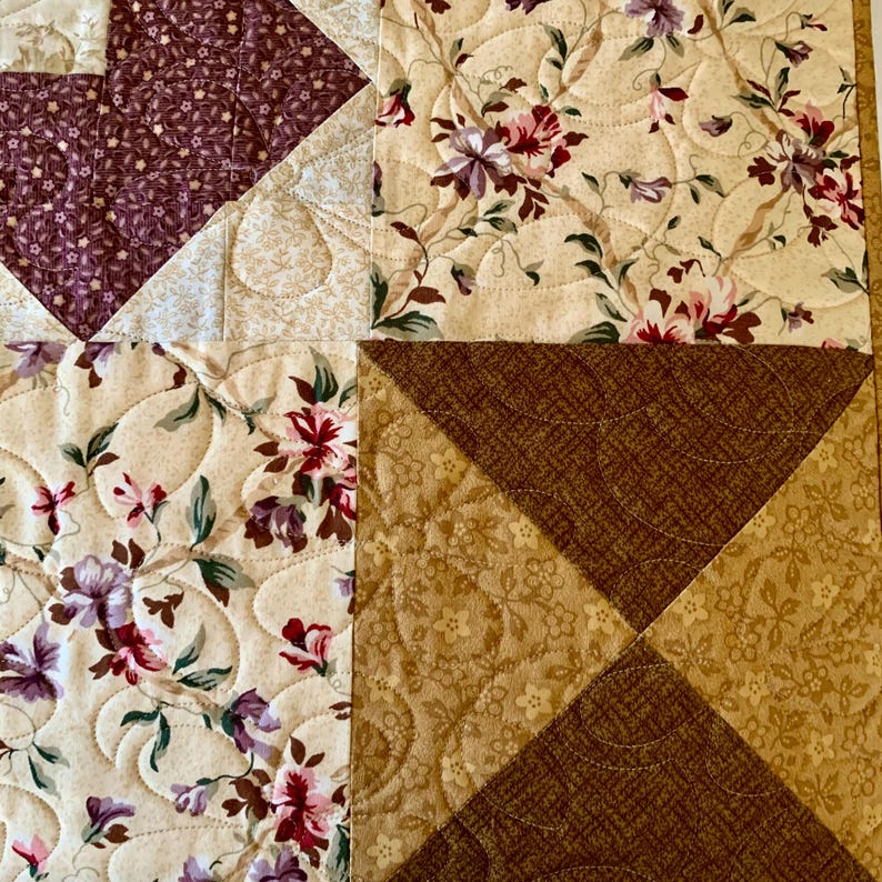 K&ouml;nnte beinhalten: Eine Nahaufnahme einer Steppdecke mit Patchwork-Muster. Die Steppdecke besteht aus verschiedenen Stoffen in Braun-, Beige- und Lilat&ouml;nen. Der Stoff hat ein Blumenmuster.