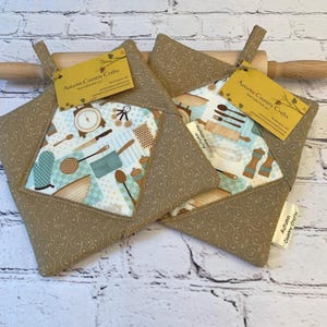 May include: Two brown and beige quilted pot holders with a kitchen utensil pattern. The pot holders have a tag that reads "Autumn Country Crafts". A wooden rolling pin is in the background.