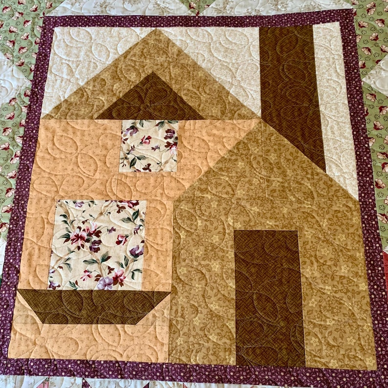 K&ouml;nnte beinhalten: Ein gequilteter Wandbehang aus Stoff mit einem braunen und beigefarbenen Hausdesign mit Blumenmustern. Das Haus hat einen Schornstein und ein Fenster. Der Stoff ist mit einem Wirbelmuster gesteppt.