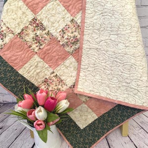 May include: A quilted blanket with a patchwork design. The quilt features squares of pink, cream, and floral fabric. The border is dark green with a floral pattern. A bouquet of pink and white tulips sits in a white pitcher.