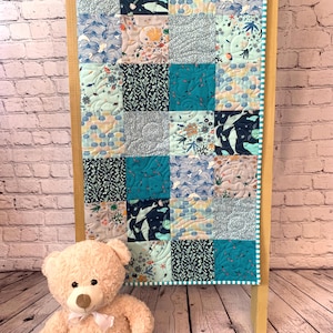 May include: A patchwork quilt with various marine-themed patterns in shades of blue, teal, and white. The quilt is displayed on a wooden frame. A beige teddy bear with a white bow sits in front of the quilt.