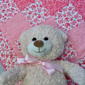 May include: A plush white teddy bear with a pink bow, sitting on a pink and white quilt with a pattern of pigs.