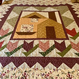 K&ouml;nnte beinhalten: Eine Quiltdecke mit Patchwork-Muster, die ein Haus in der Mitte zeigt. Die Decke ist aus verschiedenen Stoffen in Braun-, Gr&uuml;n-, Rosa- und Wei&szlig;t&ouml;nen gefertigt. Das Haus ist von einem Rand mit Tulpen und einem gewellten Saum umgeben.