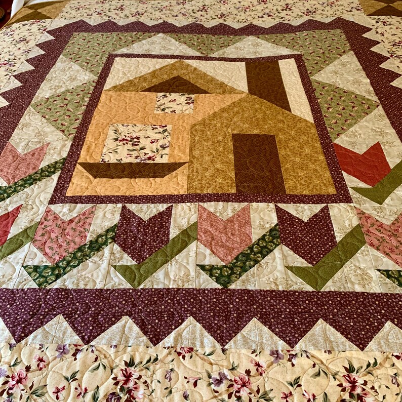 K&ouml;nnte beinhalten: Eine Quiltdecke mit Patchwork-Muster, die ein Haus in der Mitte zeigt. Die Decke ist aus verschiedenen Stoffen in Braun-, Gr&uuml;n-, Rosa- und Wei&szlig;t&ouml;nen gefertigt. Das Haus ist von einem Rand mit Tulpen und einem gewellten Saum umgeben.
