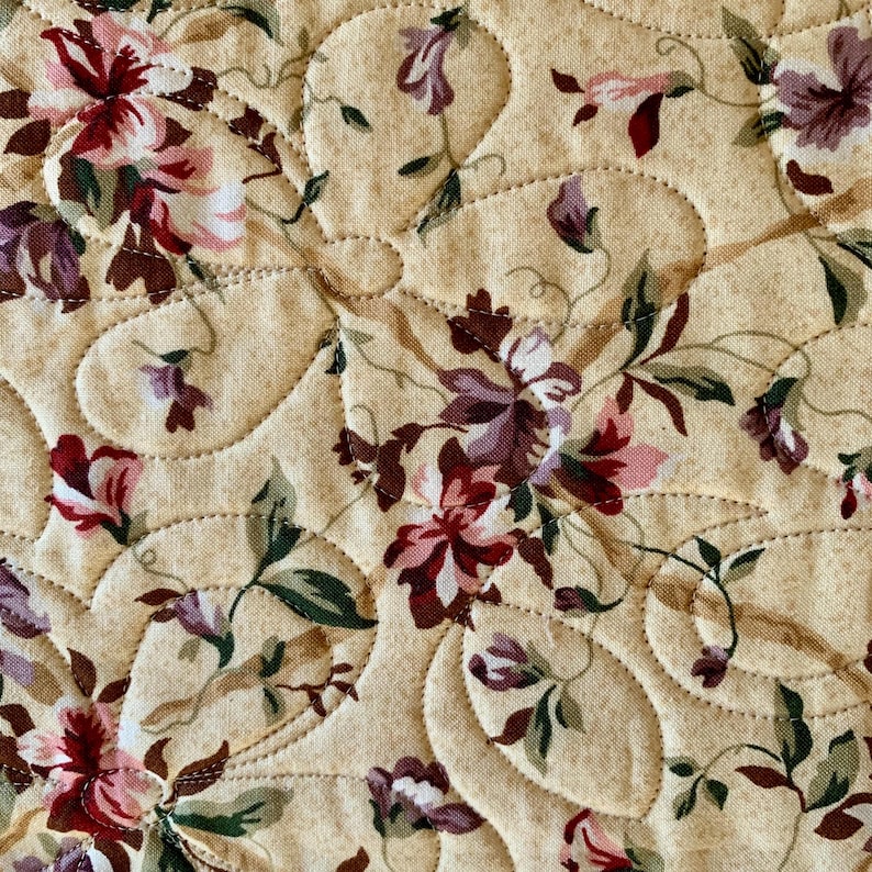 K&ouml;nnte beinhalten: Nahaufnahme eines beigefarbenen Stoffes mit gestepptem Muster. Der Stoff ist mit einem Blumendruck verziert, der rosa, rote und violette Blumen zeigt.