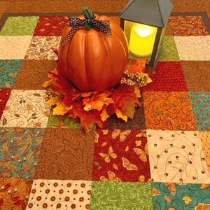 Peut inclure: Une citrouille décorative orange avec un ruban à pois noirs et blancs, entourée de feuilles d'automne et de baies artificielles, placée à côté d'une lanterne gris foncé avec une lumière jaune sur un chemin de table en tissu patchwork.