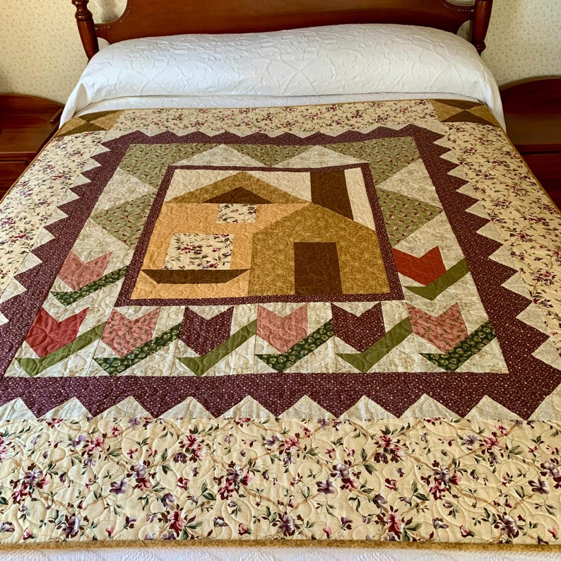 K&ouml;nnte beinhalten: Eine Quiltdecke mit einem braunen Hausmotiv in der Mitte, umgeben von einem Rand aus gr&uuml;nen und braunen Tulpen. Die Decke ist aus verschiedenen Stoffen in Braun-, Gr&uuml;n-, Rosa- und Rott&ouml;nen gefertigt.