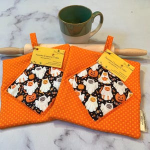 Könnte beinhalten: Zwei quadratische Topflappen aus Stoff mit orangefarbenem Punktemuster am Rand und einem schwarzen Mittelteil mit einem Motiv aus Katzen in Geisterkostümen und Kürbissen. Jeder Topflappen hat eine Aufhängeschlaufe und ein gelbes Etikett.
