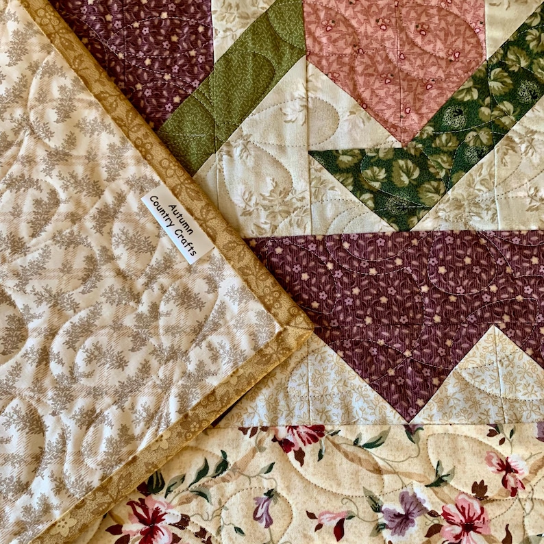 K&ouml;nnte beinhalten: Eine Nahaufnahme einer Steppdecke mit Patchwork-Muster, das verschiedene florale Muster in Rosa-, Gr&uuml;n- und Braunt&ouml;nen zeigt. Die Steppdecke ist aus verschiedenen Stoffen gefertigt und hat eine gesteppte Textur. Die Steppdecke tr&auml;gt ein Etikett mit der Aufschrift "Autumn Country Crafts".