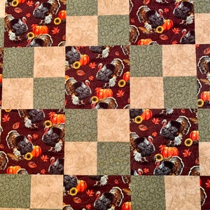 Puede incluir: Una colcha con un diseño de patchwork que presenta un patrón repetitivo de pavos, calabazas y hojas de otoño. La colcha está hecha de cuadrados de diferentes colores y patrones, incluyendo marrón, verde y beige.