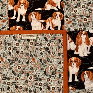 Może przedstawiać: Kołdra z czarnym tłem, na której powtarza się wzór Cavalier King Charles Spaniels. Kołdra jest obszyta tkaniną w kolorze rdzy. Kołdra ma etykietę z napisem "Autumn Country Crafts".